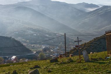 A metal cross and fence on a grassy hillside overlooking a mountain valley